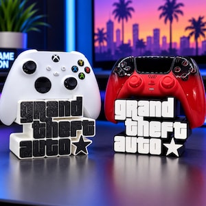 Könnte beinhalten: Ein weißer Xbox-Controller und ein rot-schwarzer PlayStation-Controller auf zwei separaten 3D-gedruckten Ständern. Jeder Ständer zeigt den Text Grand Theft Auto in einer blockartigen Schrift mit einem Sternsymbol als Halterung für Controller.