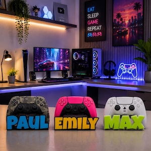 Könnte beinhalten: Drei Controller-Ständer für Videospiele mit personalisierten Namensschildern. Der linke Ständer ist tarnfarben mit blauem Schriftzug PAUL, der mittlere ist pink mit gelbem Schriftzug EMILY und der rechte ist weiß mit grünem Schriftzug MAX.