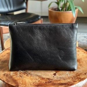 Puede incluir: Un estuche rectangular de cuero negro con un acabado liso y mate, que cuenta con una cremallera de metal plateado en el borde superior. El estuche tiene un diseño sencillo y limpio con una textura de grano natural visible en el cuero.