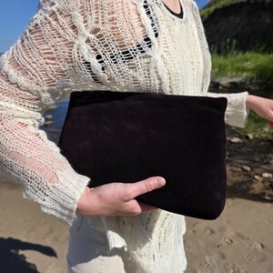 Op de afbeelding: Een rechthoekige donkerbruine suède clutch met een gladde, matte textuur. De tas heeft een eenvoudig ontwerp met strakke lijnen en een omslagsluiting, en toont een diepe, effen kleur die contrasteert met de natuurlijke omgeving buiten.