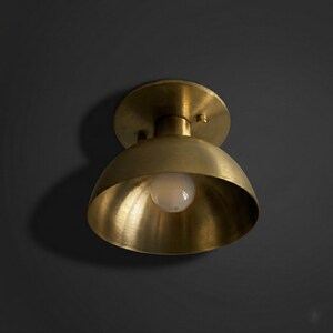 May include: A brass-colored, dome-shaped ceiling light fixture with a round base and a visible light bulb. The fixture is made of metal and has a warm, golden hue. The light is mounted on a dark gray wall.