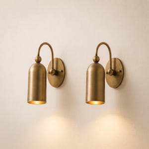 May include: A pair of matching wall sconces featuring a brushed brass finish. Each fixture consists of a circular wall plate, a curved arm, and a cylindrical shade with a rounded top. The lights emit a warm glow from the bottom of the shades.