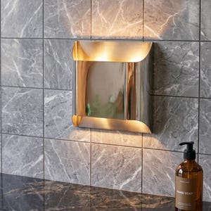May include: A modern, square wall sconce with a polished metal finish is mounted on a gray marble tile wall. The sconce emits a warm light, reflecting off the shiny surface. A brown glass bottle of hand soap sits on the countertop.