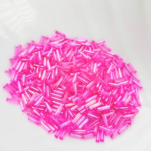 May include: A collection of bright pink glass seed beads. These small, tube-shaped beads have a translucent appearance, reflecting light. They are scattered on a white surface, creating a vivid display. Suitable for jewellery making and crafts.