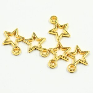 May include: A collection of gold-coloured star charms. Each charm features a hollow star shape with a small loop at the top and a circular attachment at the bottom. The charms are arranged on a white background.