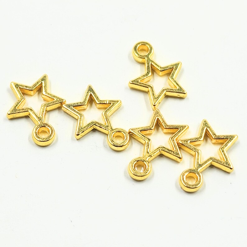 May include: A collection of gold-coloured star charms. Each charm features a hollow star shape with a small loop at the top and a circular attachment at the bottom. The charms are arranged on a white background.