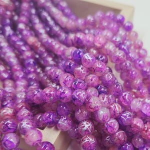 May include: Strands of purple and pink beads with a marbled effect. The beads are round and have a glossy finish. The beads are a mix of light and dark shades of purple and pink.