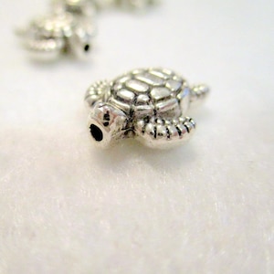 May include: A silver metal turtle charm with a detailed shell and legs. The charm has a hole through the top for attaching to a necklace or bracelet.