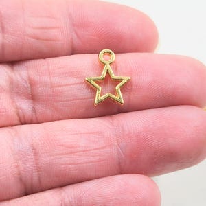 May include: A small, gold-coloured star charm with a hollow centre. The charm has a loop at the top for attaching to jewellery or other crafts. The star is held in a person's hand.