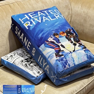 May include: Two blue rectangular cushions. One features the text "HEATED RIVALRY" and "SHANE & ILYA" with an illustration of hockey players. The other has a black and white comic strip design. Cushions are on a beige sofa.