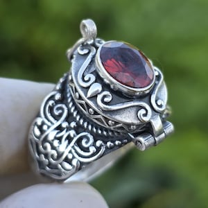 May include: A silver ring with an oval, deep red gemstone. The ring features intricate scrollwork and a hinged compartment. The gemstone is set in a decorative bezel. The ring is held up against a blurred green background.