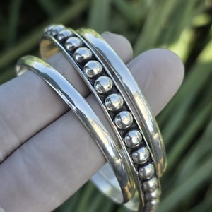 Pode incluir: Três pulseiras de prata. Uma pulseira apresenta uma fileira de pequenas contas redondas de prata. As outras duas pulseiras são lisas e polidas. As pulseiras são exibidas em um fundo verde desfocado.