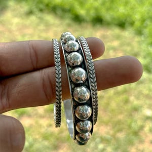 Pode incluir: Pulseiras de metal prateado. Uma pulseira apresenta uma fileira de contas grandes e redondas, ladeadas por um design trançado. A outra pulseira é uma faixa mais fina com um padrão trançado semelhante. Fundo verde desfocado.