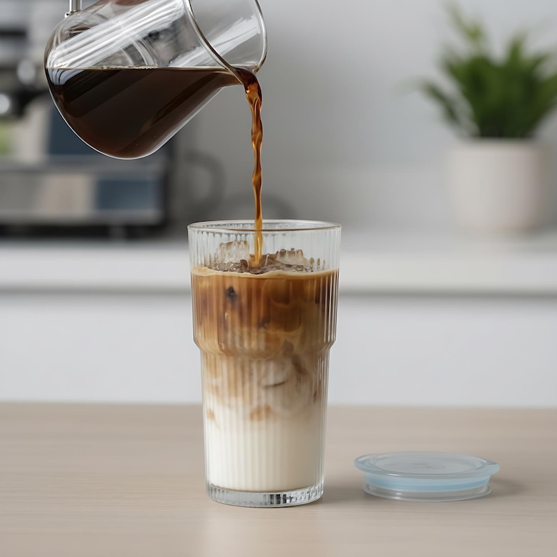 Puede incluir: Un vaso transparente lleno de hielo y leche, con caf&eacute; oscuro verti&eacute;ndose de una jarra de vidrio. El caf&eacute; crea un efecto de capas en el vaso. Una tapa azul claro est&aacute; sobre la mesa.
