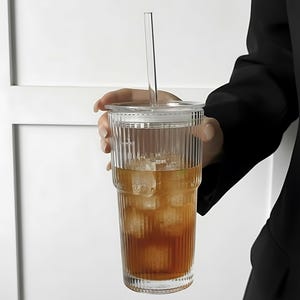 Puede incluir: Un vaso de vidrio transparente y acanalado con tapa y pajita, lleno de t&eacute; helado. El vaso lo sostiene una persona que lleva una blazer negro. La bebida es de color &aacute;mbar y los cubitos de hielo son visibles.