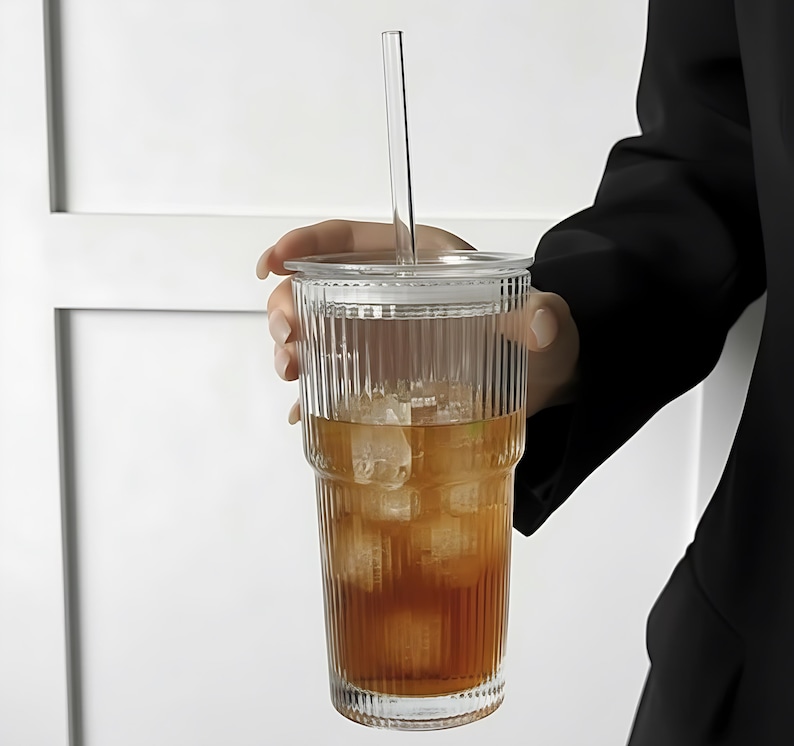 Puede incluir: Un vaso de vidrio transparente y acanalado con tapa y pajita, lleno de t&eacute; helado. El vaso lo sostiene una persona que lleva una blazer negro. La bebida es de color &aacute;mbar y los cubitos de hielo son visibles.