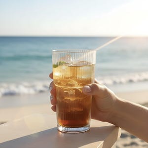 Puede incluir: Un vaso alto y transparente lleno de t&eacute; helado y cubitos de hielo, sostenido por una mano. El vaso tiene detalles verticales acanalados. La bebida es de color &aacute;mbar y se ve una ramita de menta. El fondo muestra una playa y el oc&eacute;ano.