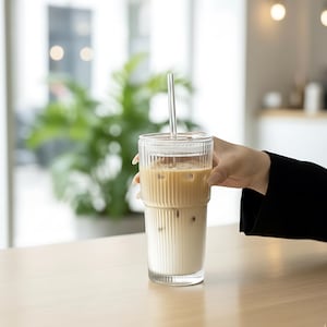 May include: A tall, clear glass with vertical ribbed detailing, filled with iced coffee and a clear straw. The glass is held by a person, and sits on a light wooden surface. The background is blurred, with a green plant visible.