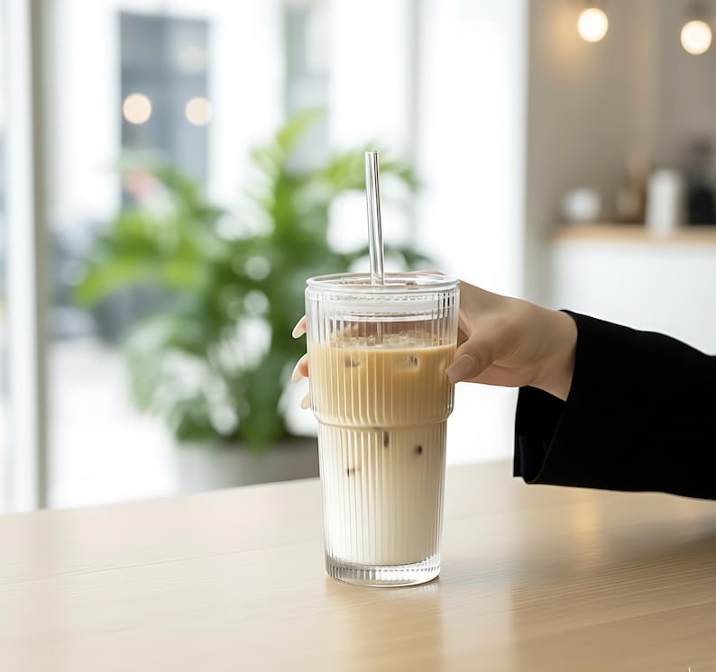 Puede incluir: Un vaso alto y transparente con detalles acanalados verticales, lleno de caf&eacute; helado y una pajita transparente. El vaso lo sostiene una persona y est&aacute; sobre una superficie de madera clara. El fondo est&aacute; borroso, con una planta verde visible.