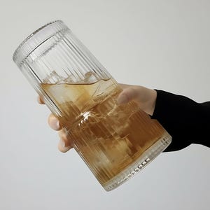 Puede incluir: Un vaso transparente y acanalado lleno de t&eacute; helado. El vaso tiene forma cil&iacute;ndrica con una superficie texturizada y lo sostiene una persona. La bebida tiene un tono marr&oacute;n dorado y se ven cubitos de hielo.