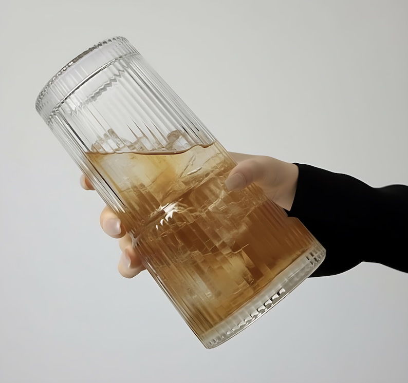 Puede incluir: Un vaso transparente y acanalado lleno de t&eacute; helado. El vaso tiene forma cil&iacute;ndrica con una superficie texturizada y lo sostiene una persona. La bebida tiene un tono marr&oacute;n dorado y se ven cubitos de hielo.