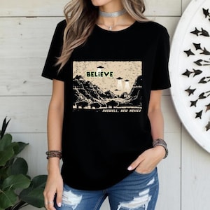 Puede incluir: Camiseta negra con un diseño gráfico en beige y negro. El diseño incluye la palabra "BELIEVE", OVNIs, un paisaje y el texto "ROSWELL, NEW MEXICO". Una prenda de vestir informal.