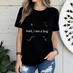 May include: Black t-shirt with the text "wait, i see a bug" and illustrations of a ladybug, ant, worm, and beetle. The shirt is worn with blue jeans.