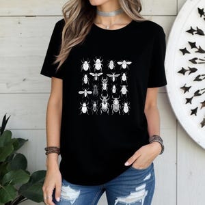 May include: Black t-shirt featuring a grid of detailed insect illustrations in white. The insects include beetles, flies, and other bugs. The shirt is a classic crew neck style and is made of a soft material.