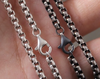 May include: Close-up of two silver-colored and two dark gray metal box chain necklaces. Each chain has a lobster clasp. The chains are made of small, rounded links. The silver chains have a bright, shiny finish, while the dark gray chains have a matte finish.