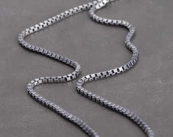 Op de afbeelding: Close-up van een zilveren box chain ketting. De ketting is gemaakt van kleine, vierkante schakels die een textuur creëren. De ketting wordt gepresenteerd op een grijze ondergrond, waarbij de ketting over het frame buigt. De sluiting is bovenaan zichtbaar.