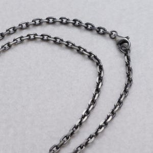 Pode incluir: Close-up de um colar de corrente prateada com fecho lagosta. Os elos da corrente são ovais e têm um acabamento texturizado e oxidado. O colar é exibido em um fundo cinza claro.
