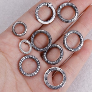 May include: A collection of silver-toned metal rings with various decorative patterns and clasp closures. Some rings feature etched designs, while others have Roman numerals or stylized text. The rings are displayed in a hand.
