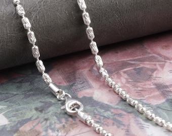 May include: A silver necklace with a detailed chain design. The chain features a combination of small, round beads and elongated, textured links. The necklace is secured with a lobster clasp.