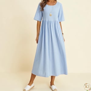 May include: A light blue midi dress with short sleeves and a gathered waist. The dress has a round neckline and falls to mid-calf. The model is wearing white slip-on shoes and a gold pendant necklace. The dress is made of a soft, flowing fabric.