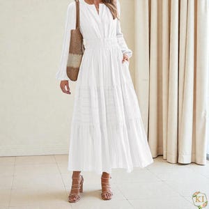 Puede incluir: Un vestido largo blanco con mangas largas, cuello en V y falda escalonada. El vestido tiene cintura ceñida y se combina con sandalias de tacón marrones y un bolso tote tejido. El vestido está hecho de una tela ligera y tiene una silueta fluida.