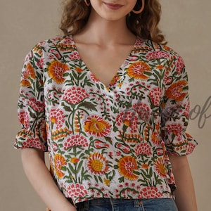 Puede incluir: Top blanco con estampado floral con cuello en V y mangas cortas. La parte superior presenta un patrón de flores naranjas y rosas con hojas verdes. Las mangas tienen un borde con volantes. La modelo lleva vaqueros azules. El texto "of Hue" es visible.