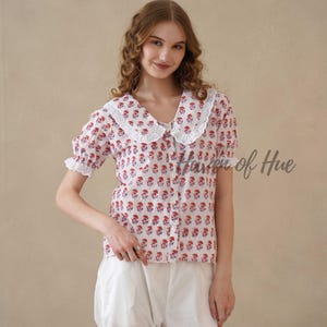 May include: A white short-sleeved blouse featuring a repeating red and blue floral block print pattern, a large scalloped white lace collar, and a front button closure. The blouse is paired with plain white trousers.