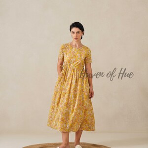 May include: A yellow floral print dress with short sleeves. The dress has a fitted bodice and a flowing skirt. The text "Haven of Hue" is visible on the right side of the image. The model is wearing white sandals and standing on a round jute rug.