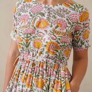 May include: White floral print dress with short sleeves and a drawstring waist. The dress features a pattern of pink and yellow flowers with green leaves. The dress has pockets and a round neckline.