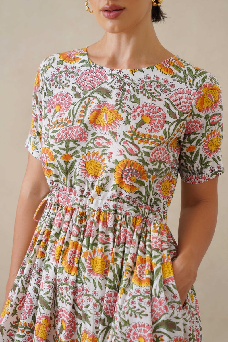 May include: White floral print dress with short sleeves and a drawstring waist. The dress features a pattern of pink and yellow flowers with green leaves. The dress has pockets and a round neckline.