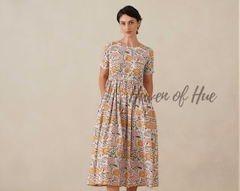 May include: A midi-length dress featuring a white base with a repeating floral pattern in shades of yellow, pink, and green. The dress has short sleeves, a round neckline, a gathered waistline, and a full skirt. The text "Woven of Hue" is visible.