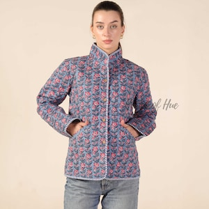 May include: A quilted jacket in a muted blue hue featuring a repeating floral pattern in shades of pink and dark green. The garment has a high collar, long sleeves, two front pockets, and a button-down front closure with white and blue striped trim.