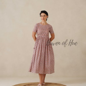 May include: A light pink midi dress with a floral pattern. The dress has short sleeves and a cinched waist. The text "Power of Hue" is visible on the right side of the image. The person is standing on a round, woven rug.