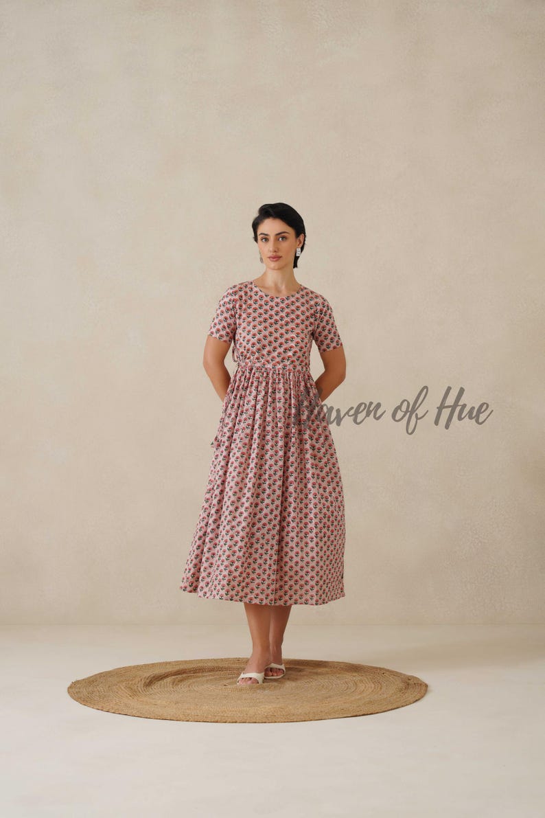 May include: A light pink midi dress with a floral pattern. The dress has short sleeves and a cinched waist. The text "Power of Hue" is visible on the right side of the image. The person is standing on a round, woven rug.