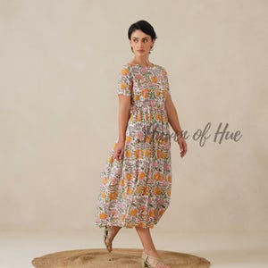 May include: A white, short-sleeved dress with a floral pattern in shades of yellow, pink, and green. The dress has a round neckline and a gathered waist. The text "Vision of Hue" is visible on the right side of the image. The model is wearing green sandals.