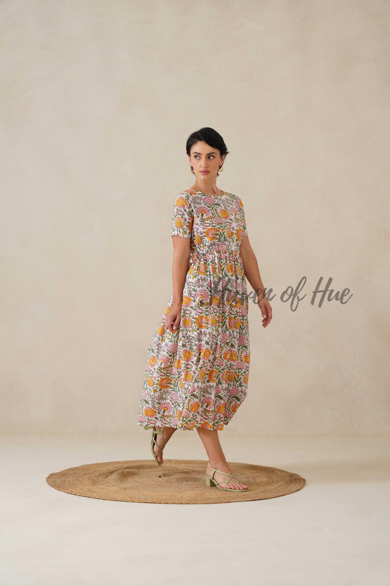 May include: A white, short-sleeved dress with a floral pattern in shades of yellow, pink, and green. The dress has a round neckline and a gathered waist. The text "Vision of Hue" is visible on the right side of the image. The model is wearing green sandals.