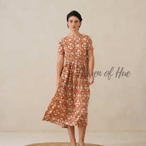 May include: A woman wearing a red midi dress with a repeating floral pattern in white and green. The dress has short sleeves and a gathered waist. The text "even of Hue" is visible on the right side of the image. The woman is wearing gold sandals.