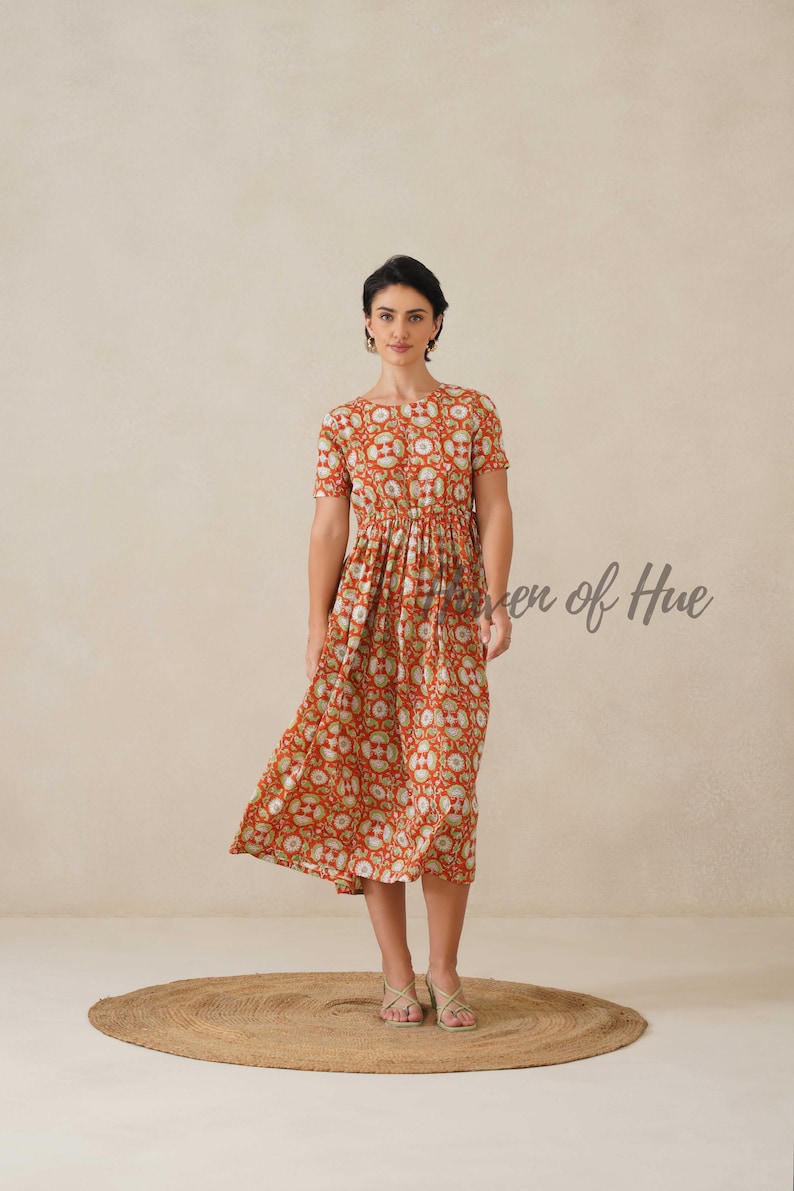 May include: A woman wearing a red midi dress with a repeating floral pattern in white and green. The dress has short sleeves and a gathered waist. The text "even of Hue" is visible on the right side of the image. The woman is wearing gold sandals.