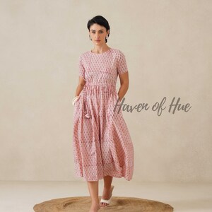 May include: A light pink, short-sleeved dress with a floral pattern. The dress has a gathered waist and pockets. The text "Haven of Hue" is visible on the right side. The model is wearing white sandals.