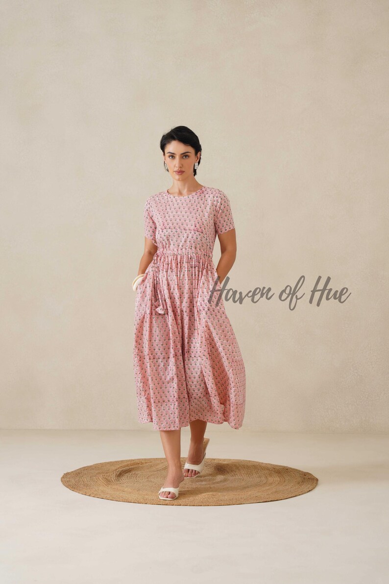 May include: A light pink, short-sleeved dress with a floral pattern. The dress has a gathered waist and pockets. The text "Haven of Hue" is visible on the right side. The model is wearing white sandals.